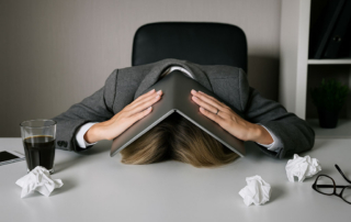 Erschöpfte Frau im Büro mit Laptop über dem Kopf – Symbolbild für Burnout und Überlastung